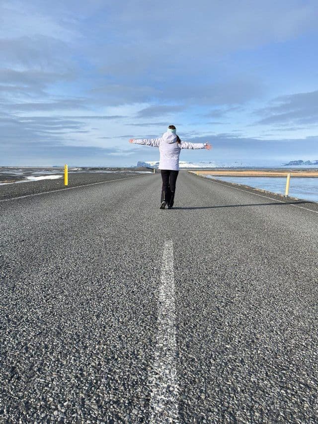 Una persona vista di spalle cammina con le braccia distese al centro di una strada vuota in un vasto paesaggio piatto e innevato.