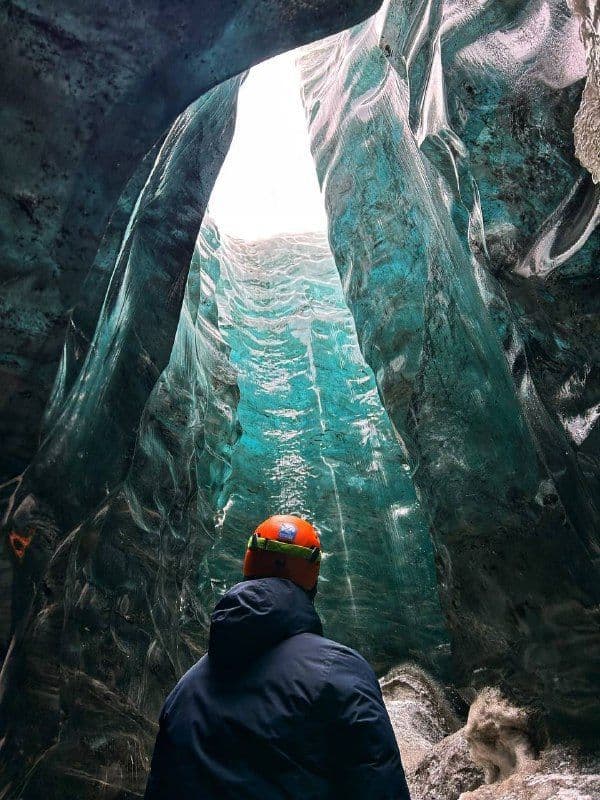 Una persona con un casco arancione si trova all'interno di una grotta di ghiaccio blu, guardando verso un'apertura nel cielo.