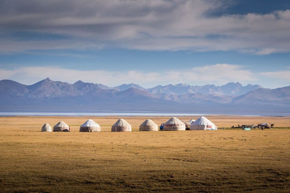 A row of yurts on a grassy plain in front of a lake and a distant mountain range.