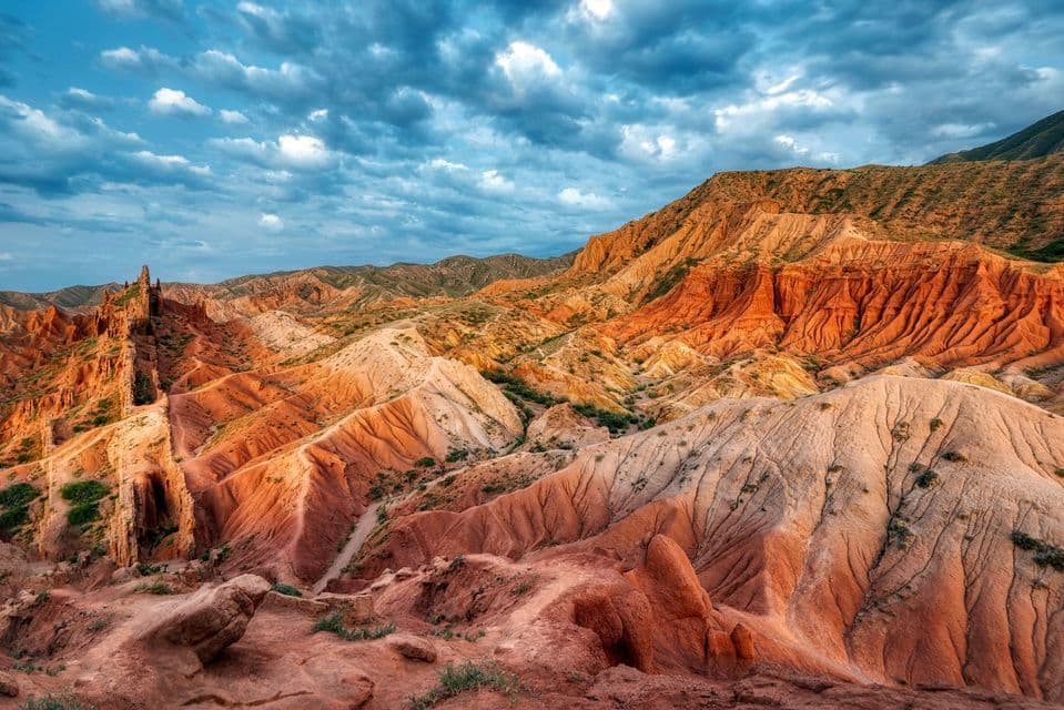 A wide view of an arid landscape of red and orange canyon formations under a cloudy blue sky.