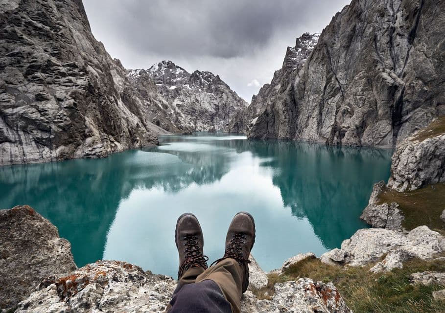 Una vista en primera persona de botas de senderismo en una cornisa rocosa, con vistas a un lago tranquilo de color turquesa rodeado de montañas empinadas bajo un cielo nublado.