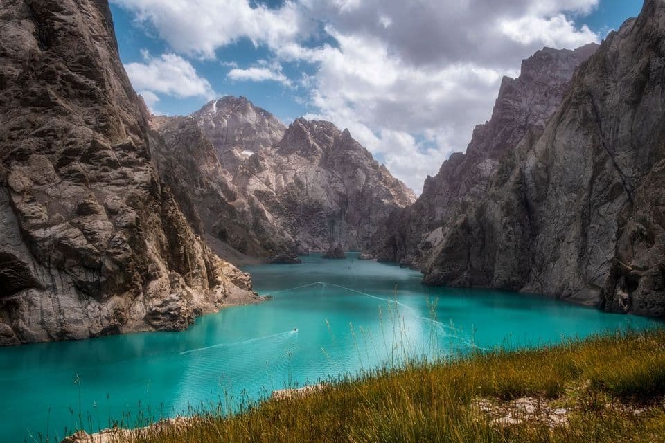 Un petit bateau laisse un sillage sur un lac turquoise, entouré de montagnes rocheuses escarpées, sous un ciel nuageux.