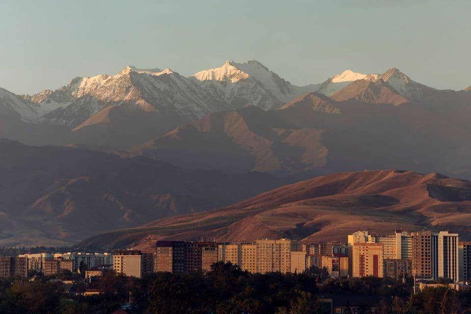 El horizonte de una ciudad se alza al pie de colinas onduladas, con una gran cordillera nevada de fondo.