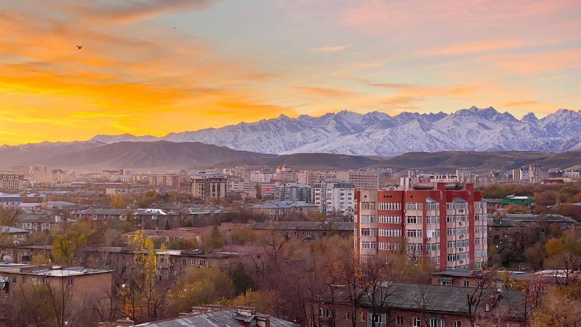 Eine Stadtlandschaft mit Gebäuden vor einer Kette schneebedeckter Berge unter einem goldenen Sonnenuntergangshimmel.