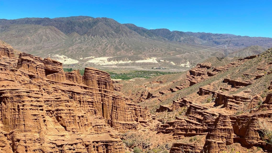 Une vue sur un canyon avec des formations rocheuses rouges stratifiées, et des montagnes arides au loin sous un ciel bleu vif.