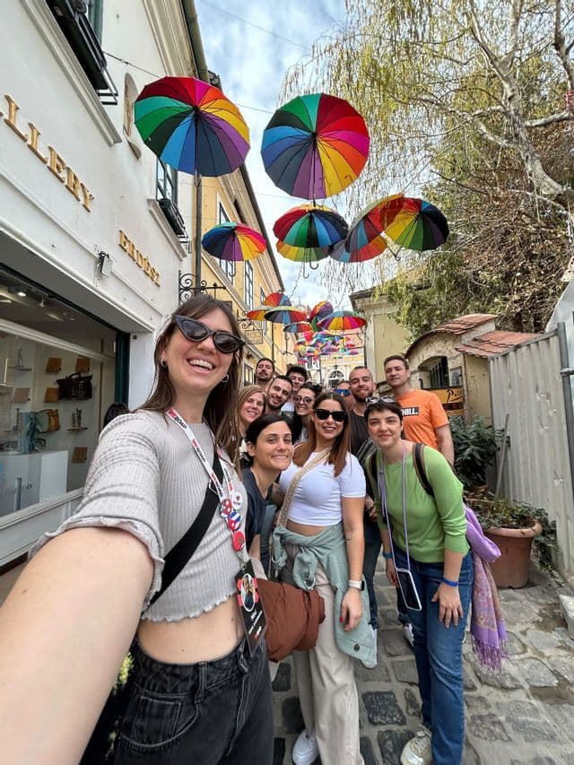 Un grupo de WeRoad posa para una selfie en una calle adoquinada bajo un dosel de coloridos paraguas arcoíris.