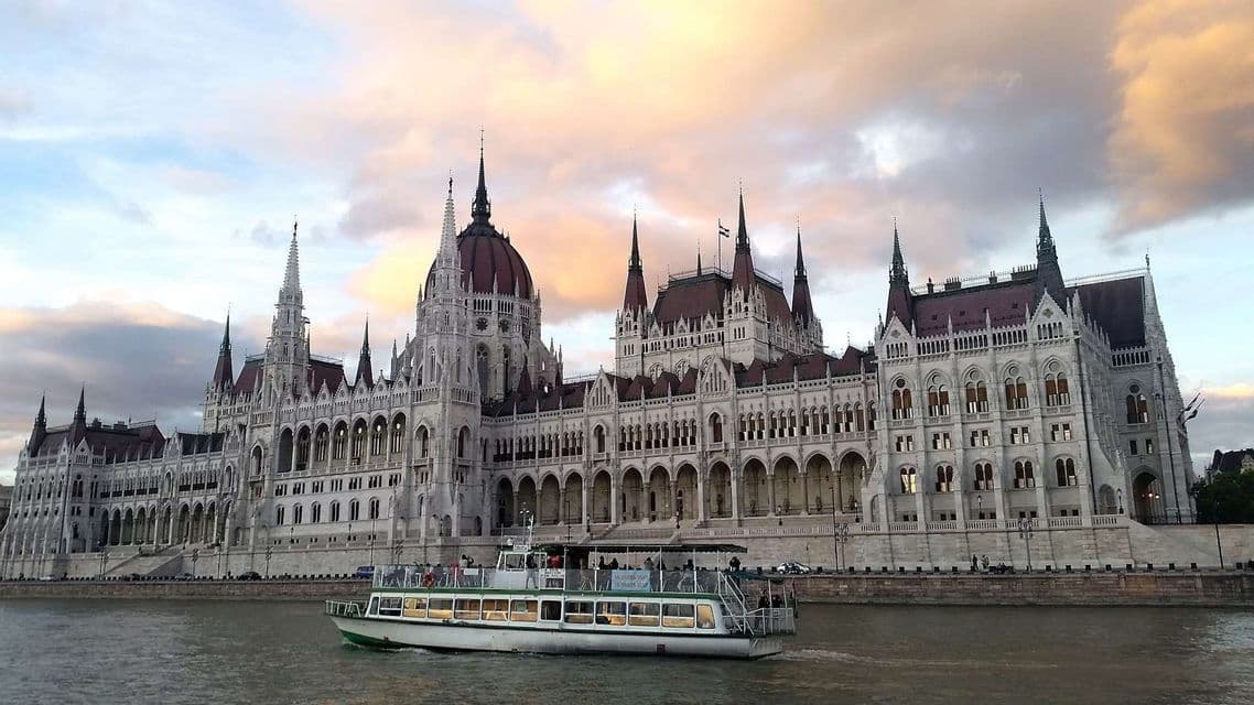 Un barco turístico con pasajeros navega por un río frente a un edificio masivo y ornamentado con cúpulas y agujas bajo un cielo nublado.