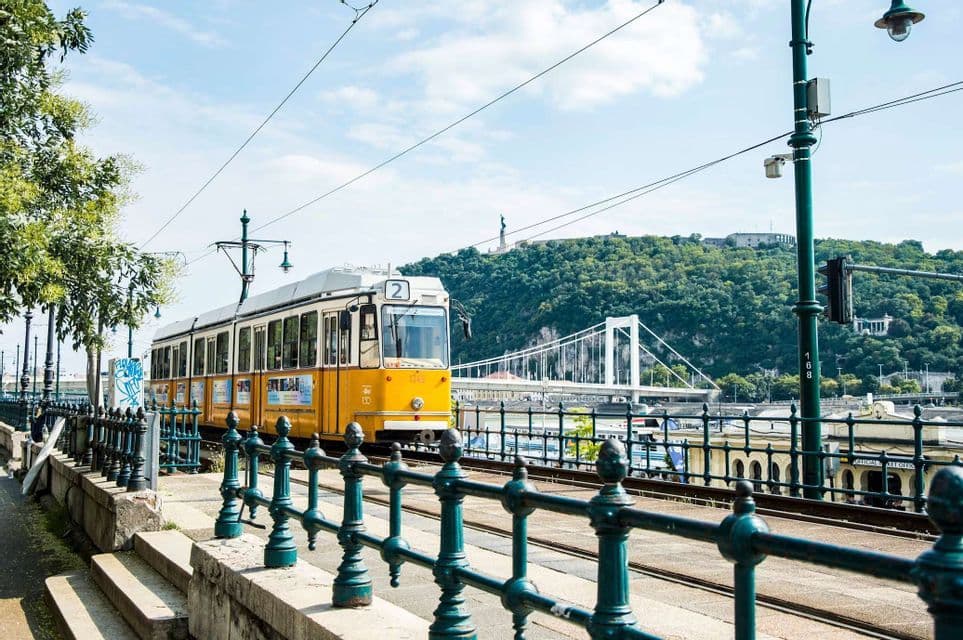 Un tranvía amarillo en sus vías junto a un andén de piedra, con un puente blanco y una colina verde al fondo.