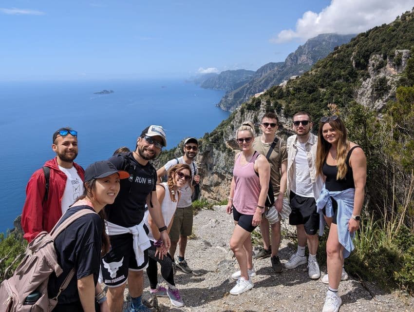 Eine WeRoad-Gruppenreise posiert für ein Foto auf einem Wanderweg mit Blick auf eine bergige Küste und das blaue Meer.