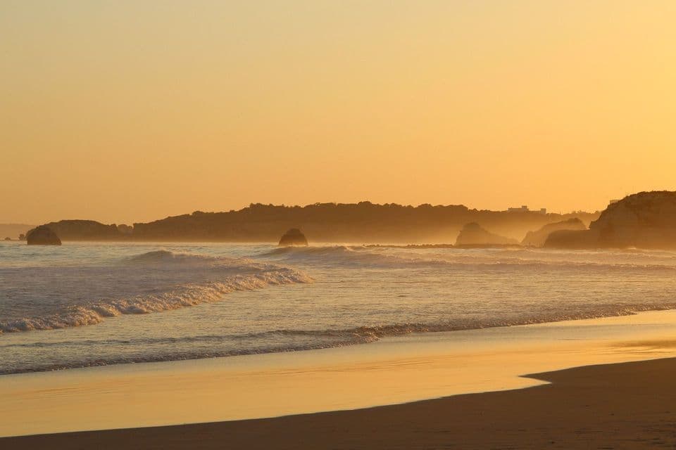 Le onde dell'oceano si infrangono su una spiaggia sabbiosa durante un tramonto dorato, con scogliere nebbiose visibili lungo la costa distante.