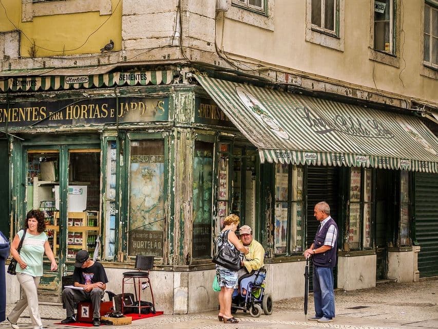 Gente su un marciapiede di città, davanti a una bottega d'angolo rustica e vissuta, con una tenda a strisce verdi e bianche.