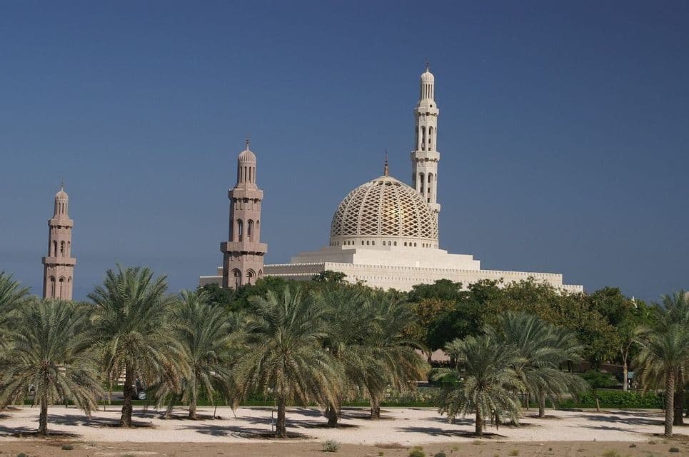Una grande moschea bianca con una cupola dorata a traliccio e minareti si erge dietro un boschetto di palme sotto un cielo azzurro chiaro.