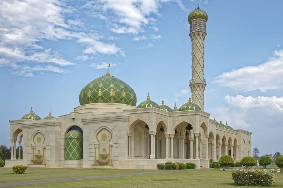 Una moschea riccamente decorata con cupole verdi e dorate e un alto minareto si erge su un prato ben curato sotto un cielo parzialmente nuvoloso.