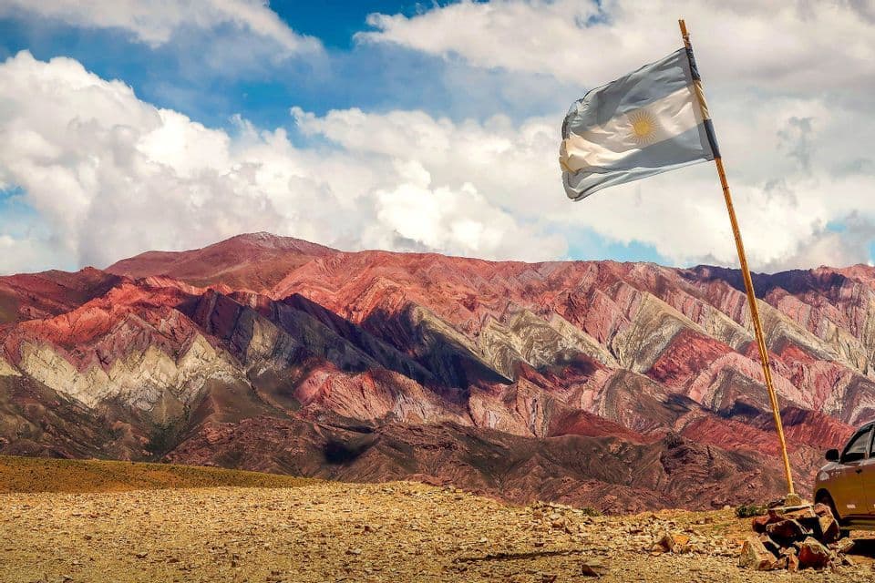 Bandiera argentina sventola davanti a una catena montuosa colorata e stratificata sotto un cielo parzialmente nuvoloso.