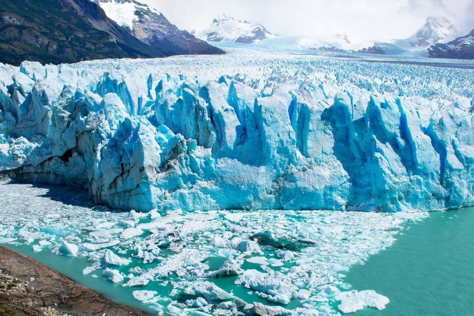 Una maestosa e scolpita parete di ghiacciaio blu che incontra un lago turchese, costellato di iceberg galleggianti, con montagne innevate sullo sfondo.