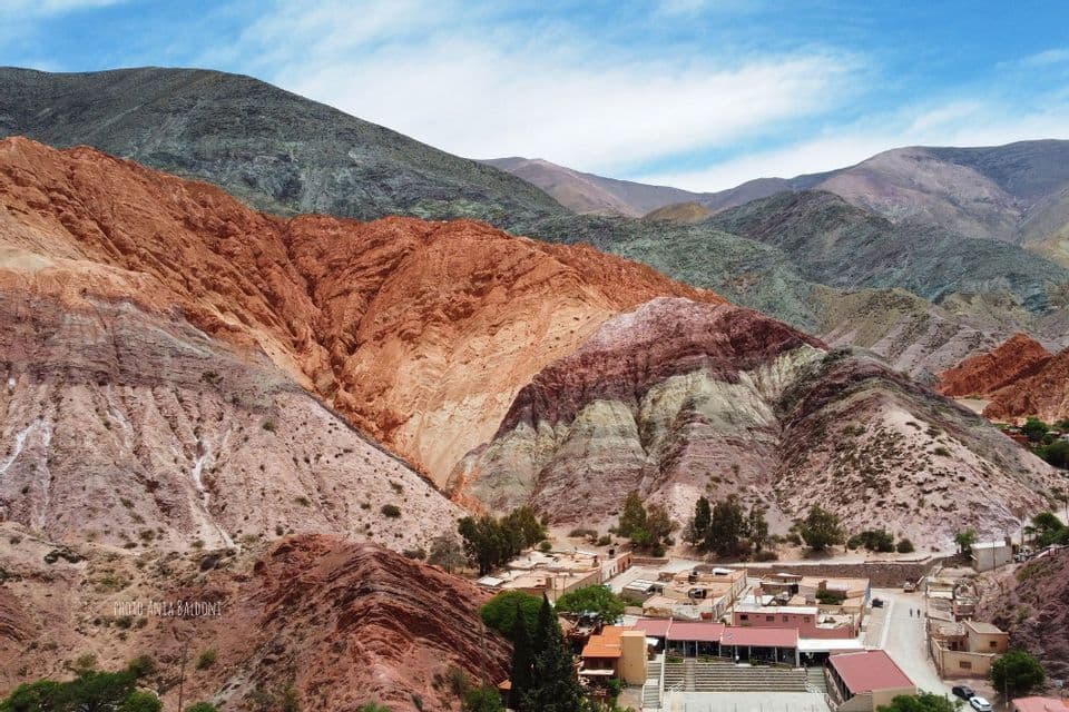 Un piccolo villaggio con edifici bassi e alberi adagiato ai piedi di grandi montagne colorate con strati di roccia rossa, arancione e verde.