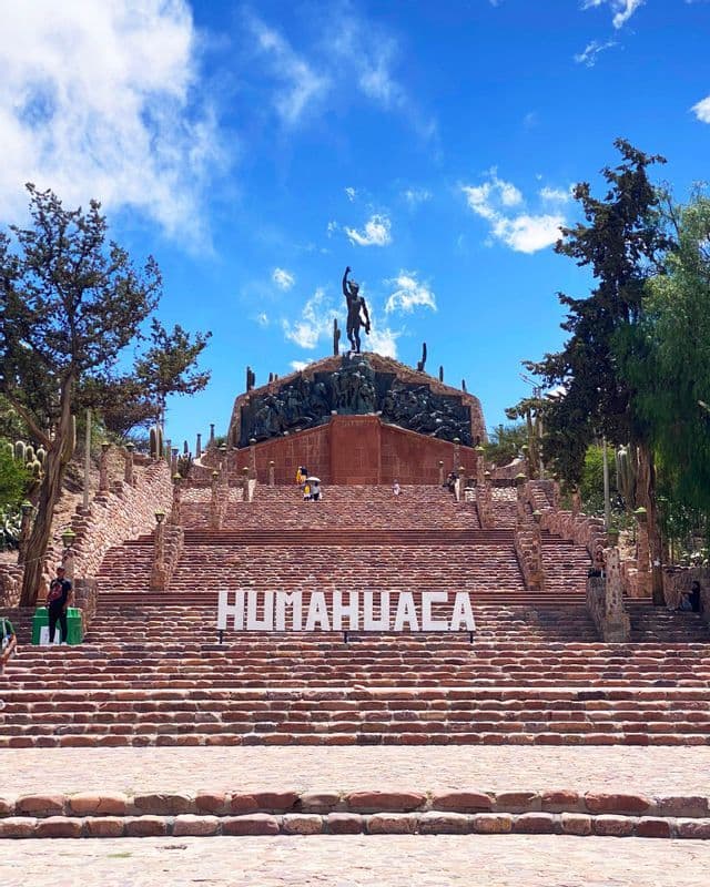 L'insegna bianca 'HUMAHUACA' si trova su un'ampia scalinata in pietra che porta a un monumento, sotto un cielo azzurro con qualche nuvola.
