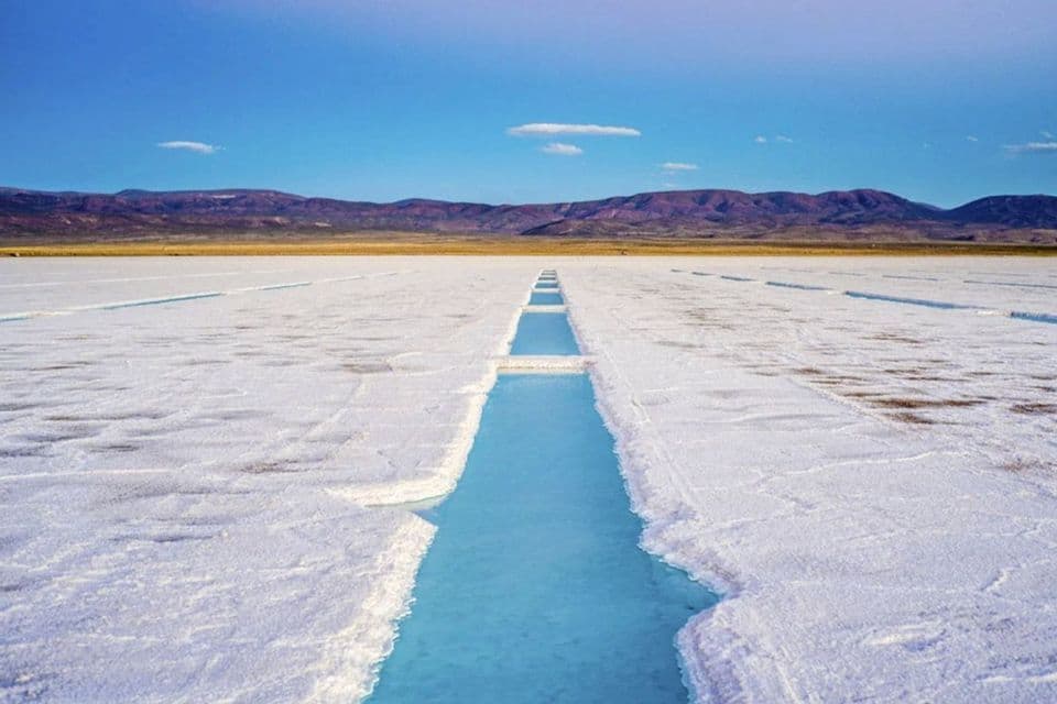 Canali di acqua turchese che solcano una vasta distesa di sale bianco con montagne lontane sotto un cielo azzurro e limpido.