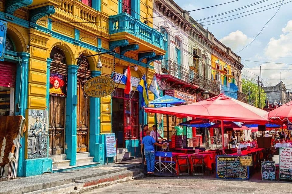 Una strada colorata con edifici dipinti di giallo brillante e blu, e un caffè all'aperto con tavoli e ombrelloni rossi.