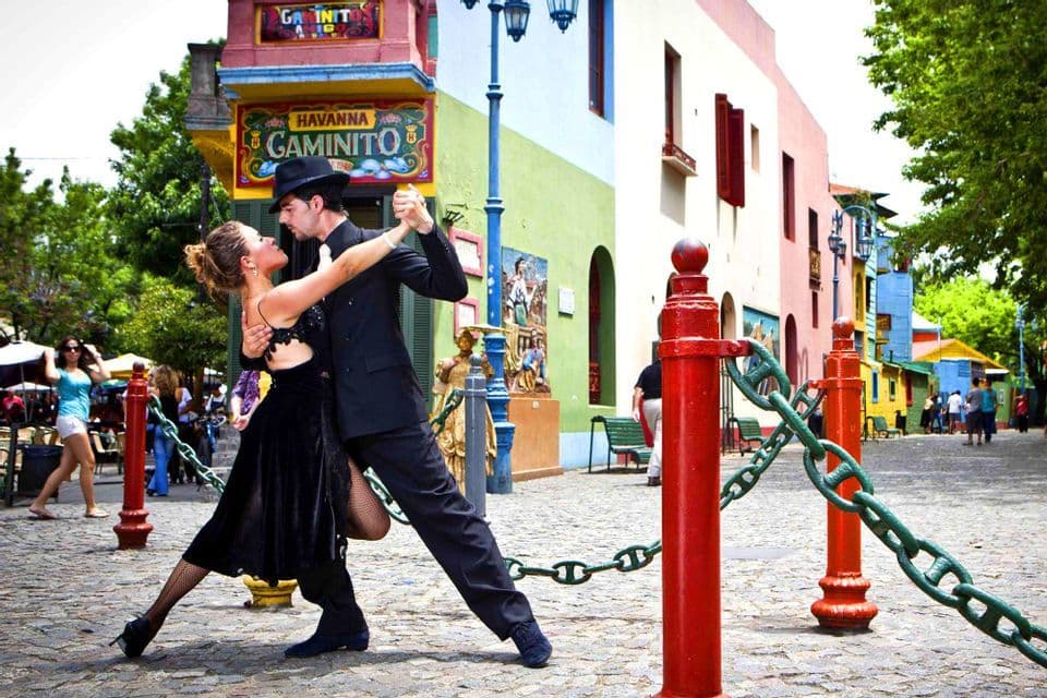 Un uomo e una donna vestiti di nero ballano il tango su una strada di ciottoli fiancheggiata da edifici colorati.