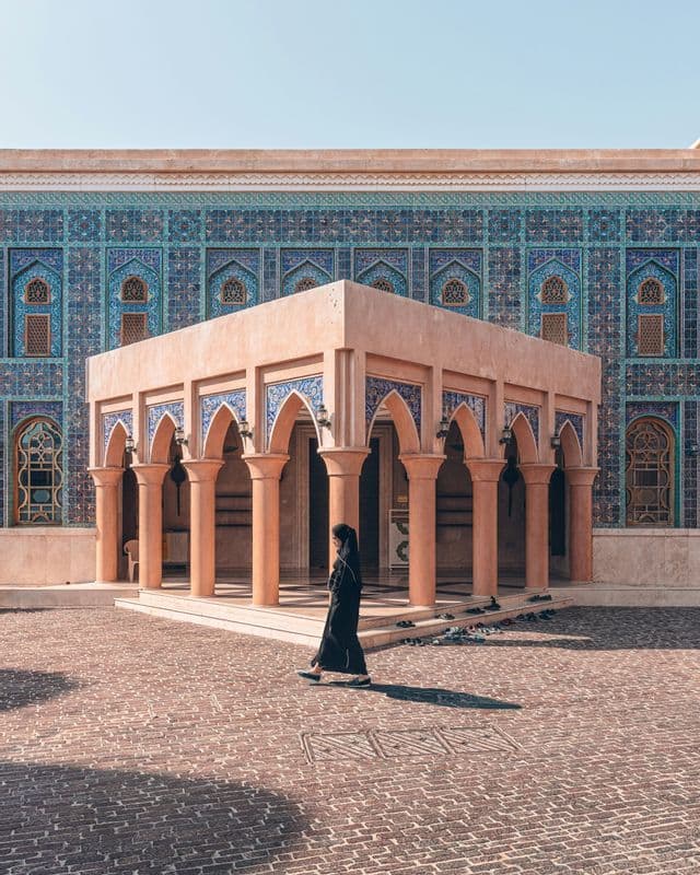 Una donna in abaya nera attraversa un cortile pavimentato di fronte a un edificio con elaborate piastrelle a mosaico blu.
