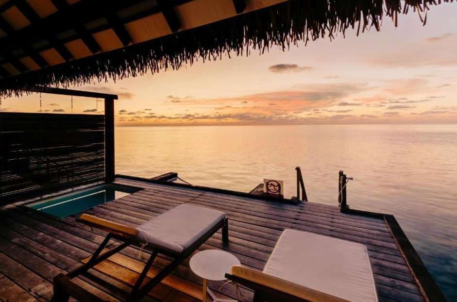 Deux chaises longues et une petite piscine sur une terrasse en bois d'une villa sur pilotis, avec vue sur un océan calme au coucher du soleil.