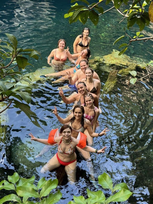 Eine WeRoad-Gruppenreise posiert in einer Reihe auf einem untergetauchten Baumstamm in klarem türkisfarbenem Wasser, umgeben von Dschungelbewuchs.