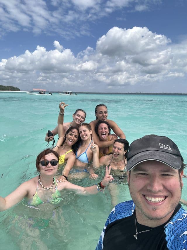 Eine WeRoad-Gruppenreise macht ein Gruppenselfie beim Schwimmen in klarem türkisfarbenem Wasser unter einem teilweise bewölkten Himmel.