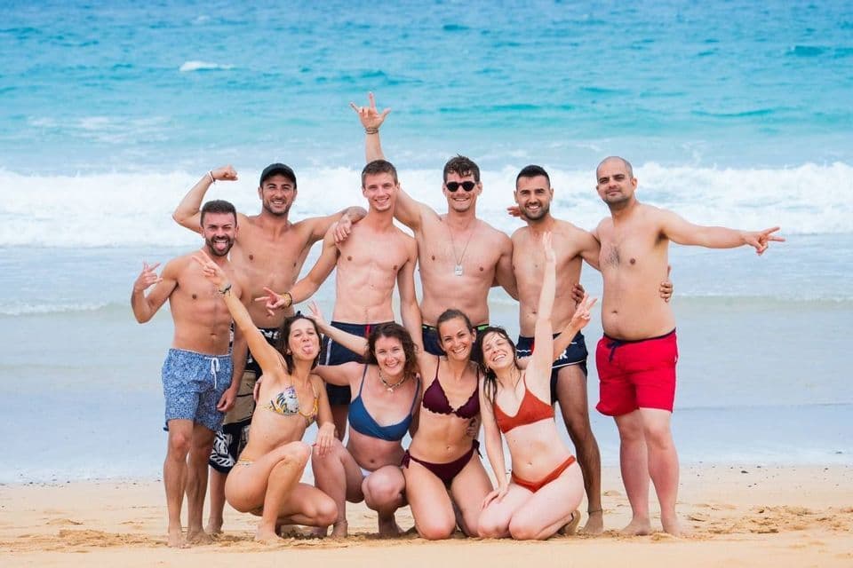 Un viaje en grupo de WeRoad de hombres y mujeres en traje de baño posando y sonriendo para una foto en una playa de arena.