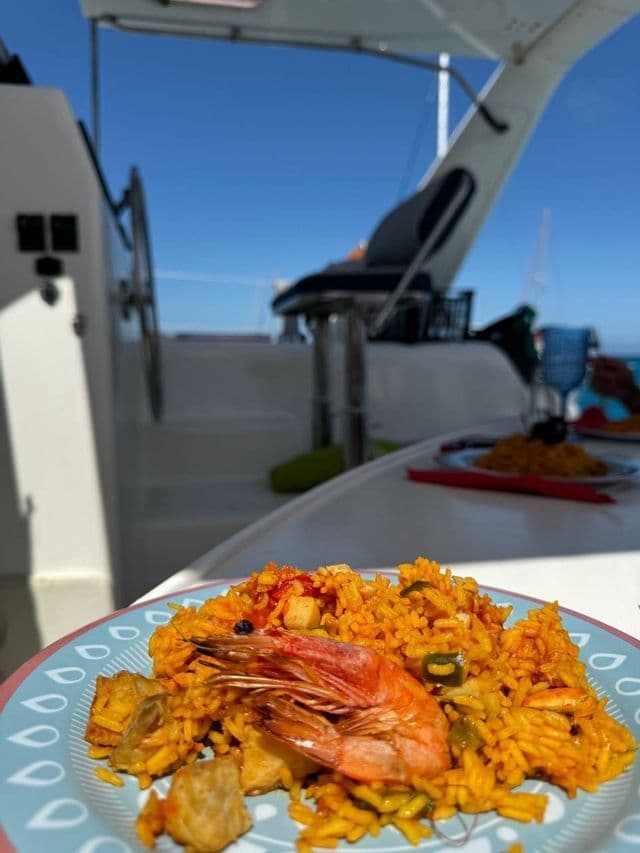 Un primo piano di un piatto di paella con un gamberone, servita sul ponte di una barca sotto un cielo soleggiato.