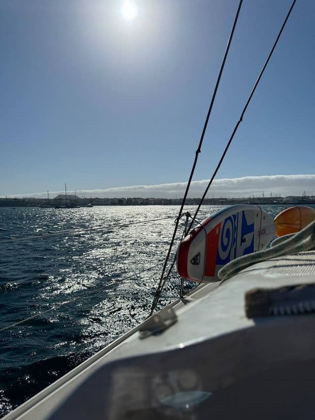 Un paddleboard è attaccato a una barca a vela su acque illuminate dal sole, con un litorale lontano sotto un sole splendente.