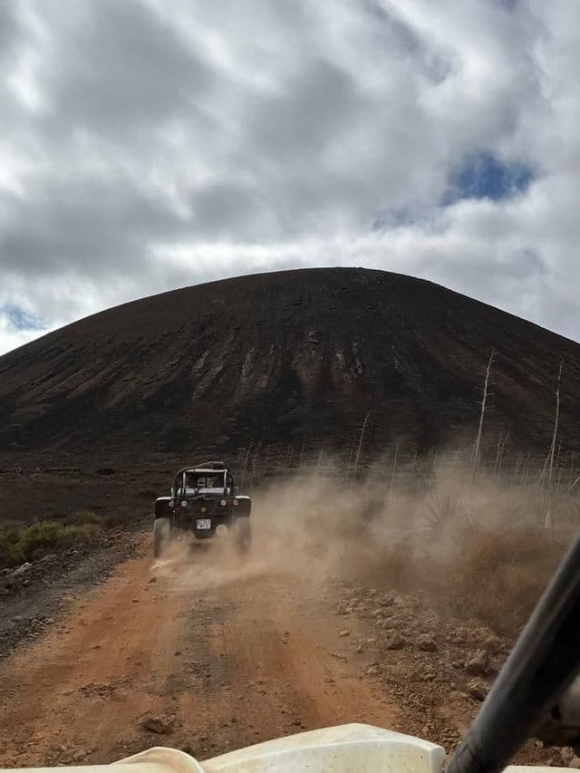 Un vehículo todoterreno levanta polvo mientras avanza por un camino de tierra hacia una montaña grande y oscura bajo un cielo nublado.
