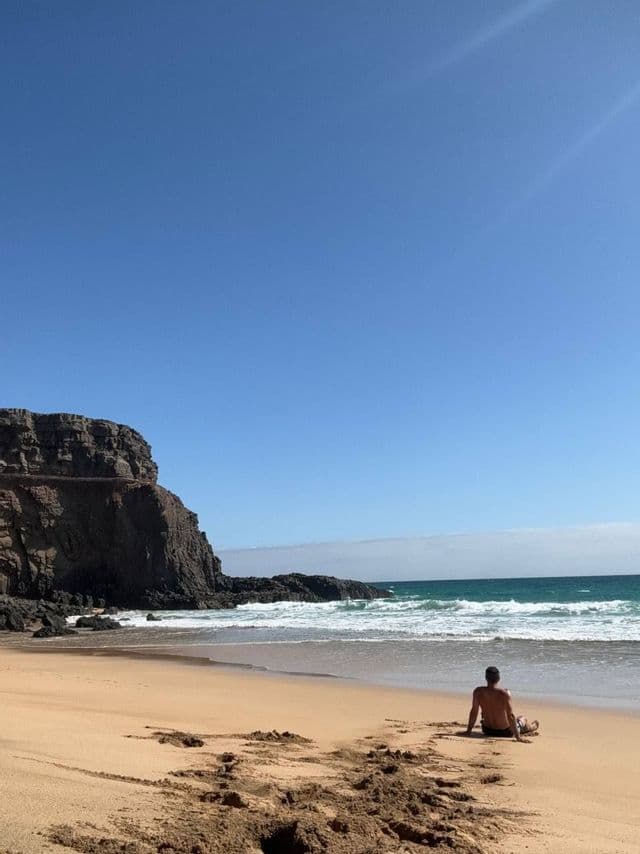 Una persona se sienta en una playa de arena frente al océano, con un gran acantilado rocoso a la izquierda bajo un cielo despejado.