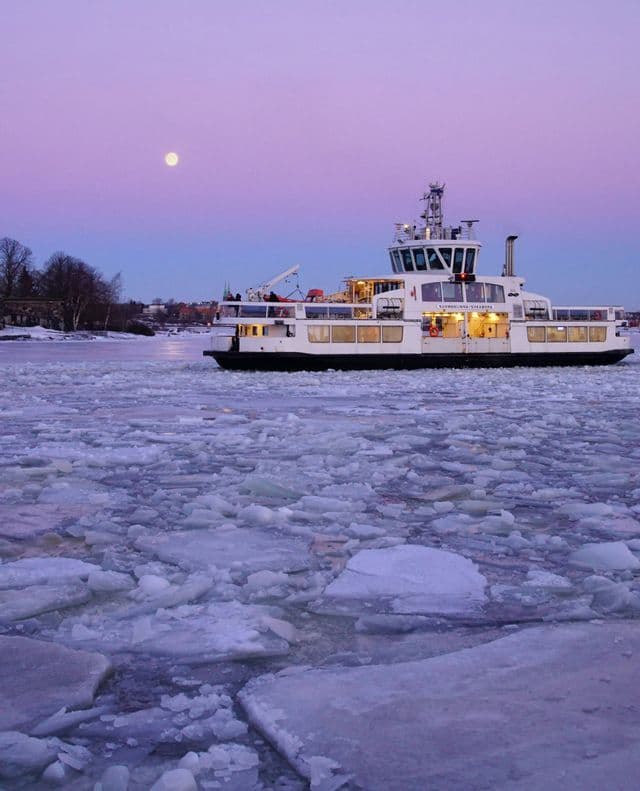 Un traghetto bianco naviga attraverso un mare di ghiaccio sotto una luna piena e un cielo crepuscolare viola.