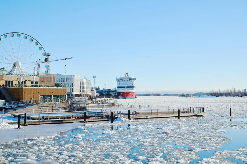 Un porto cittadino in una giornata di sole con un mare ghiacciato, un traghetto ormeggiato, una piscina all'aperto e una grande ruota panoramica.