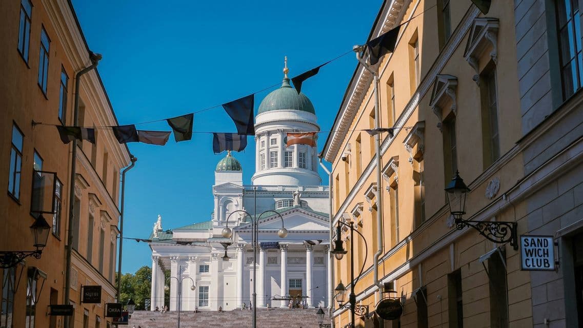 Una cattedrale bianca con una cupola verde è incorniciata da edifici gialli su una strada stretta con bandiere appese sotto un cielo azzurro chiaro.