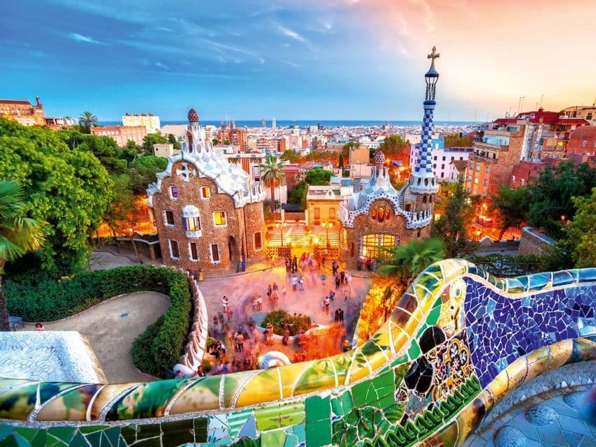 Una vista sopraelevata su una panchina a mosaico colorata di due edifici unici in una piazza al tramonto, con uno skyline cittadino e il mare sullo sfondo.