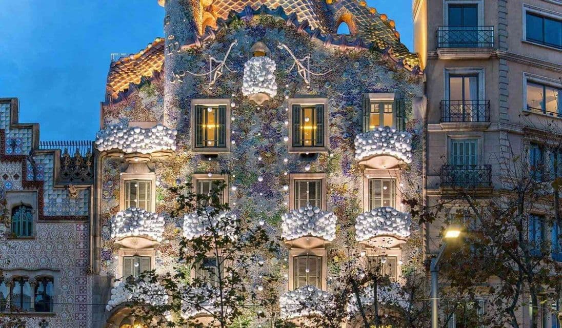 Un edificio con una facciata a mosaico colorata, balconi arrotondati unici e un tetto ad arco a squame è illuminato al tramonto.