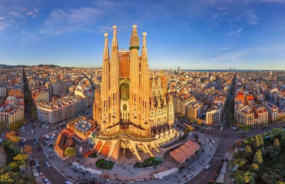 Una vista aerea e panoramica di una grande cattedrale con molte guglie al centro di una fitta città sotto un cielo blu.
