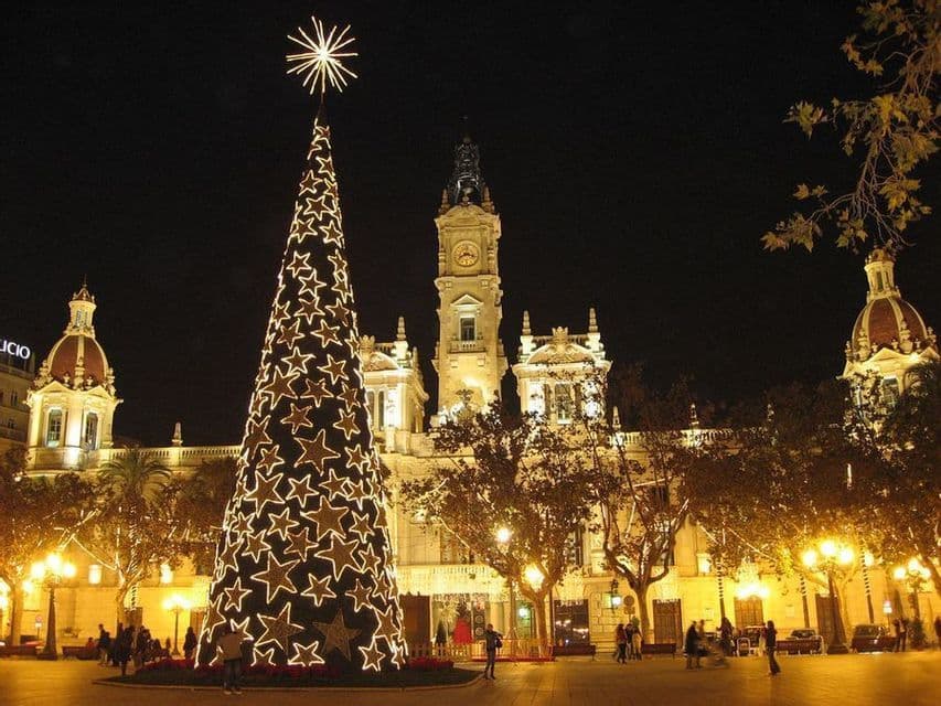 Un albero di Natale illuminato e decorato con stelle splendenti si erge in una piazza cittadina di notte, di fronte a un grande e sontuoso edificio.
