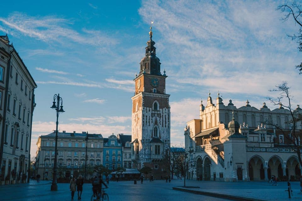 Una alta y histórica torre del reloj se alza en el centro de una gran plaza adoquinada, rodeada de edificios antiguos bajo un cielo azul.