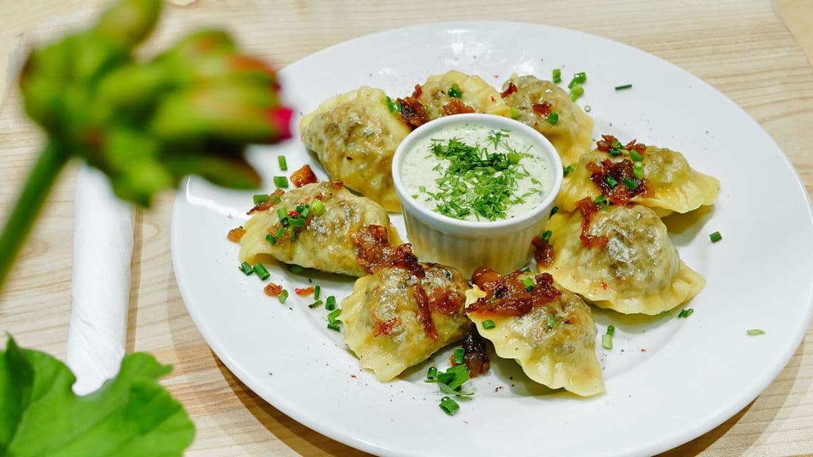 Pierogi en un plato blanco, cubiertos con cebolla frita y cebollino, con un tazón central de salsa cremosa de hierbas.