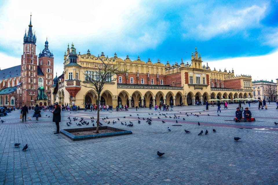 Una gran plaza empedrada llena de gente y palomas, con ornamentados edificios históricos de fondo bajo un cielo azul.