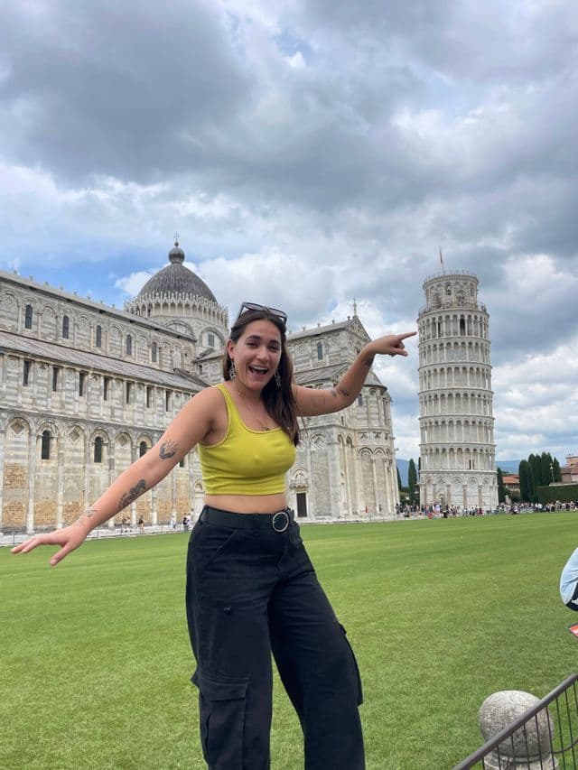 Eine Frau in einem gelben Oberteil lächelt und zeigt auf den Schiefen Turm von Pisa auf einem grünen Rasen unter bewölktem Himmel.