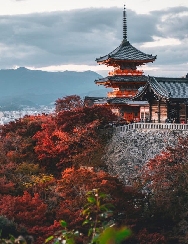 Eine mehrstufige japanische Pagode steht auf einem von rotem Herbstlaub bedeckten Hügel, mit Bergen im Hintergrund.