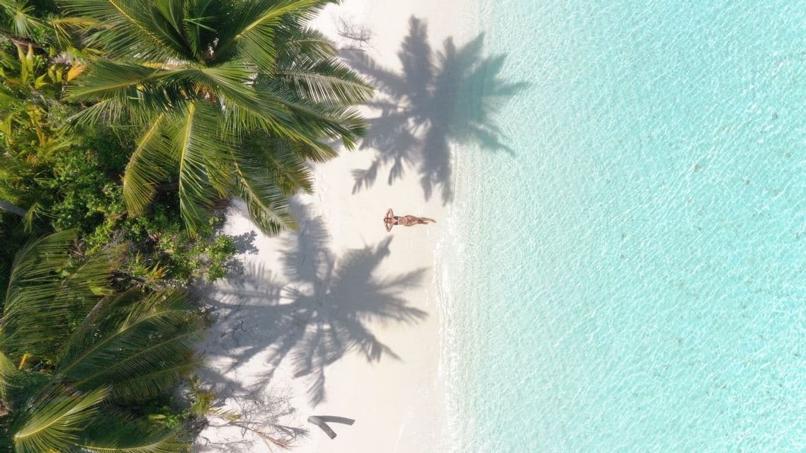 Vista aérea de una mujer relajándose en una playa de arena blanca, con sombras de palmeras y agua turquesa cristalina.