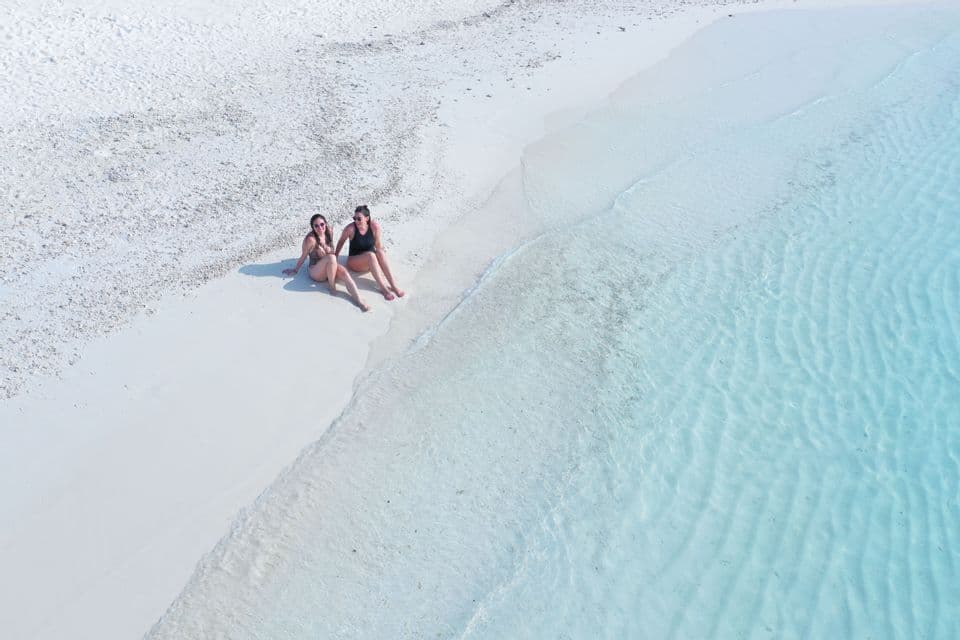 Vista aérea de dos mujeres en traje de baño sentadas en una playa de arena blanca al borde del agua turquesa cristalina.
