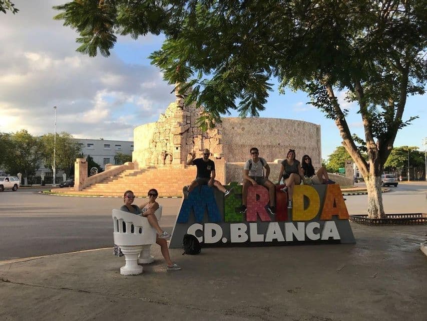 Eine WeRoad-Gruppe posiert vor dem farbenfrohen Merida-Stadtschild, im Hintergrund ein großes Steinmonument.