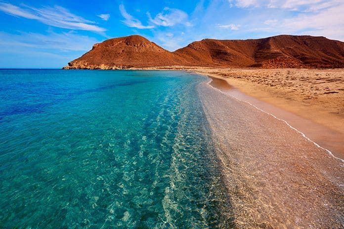 La riva di una spiaggia sabbiosa con acqua turchese limpida e poco profonda e colline aride sullo sfondo.
