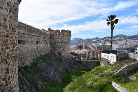 Una cinta muraria in pietra con torri si erge su una collina erbosa che domina una moderna città costiera e una palma.